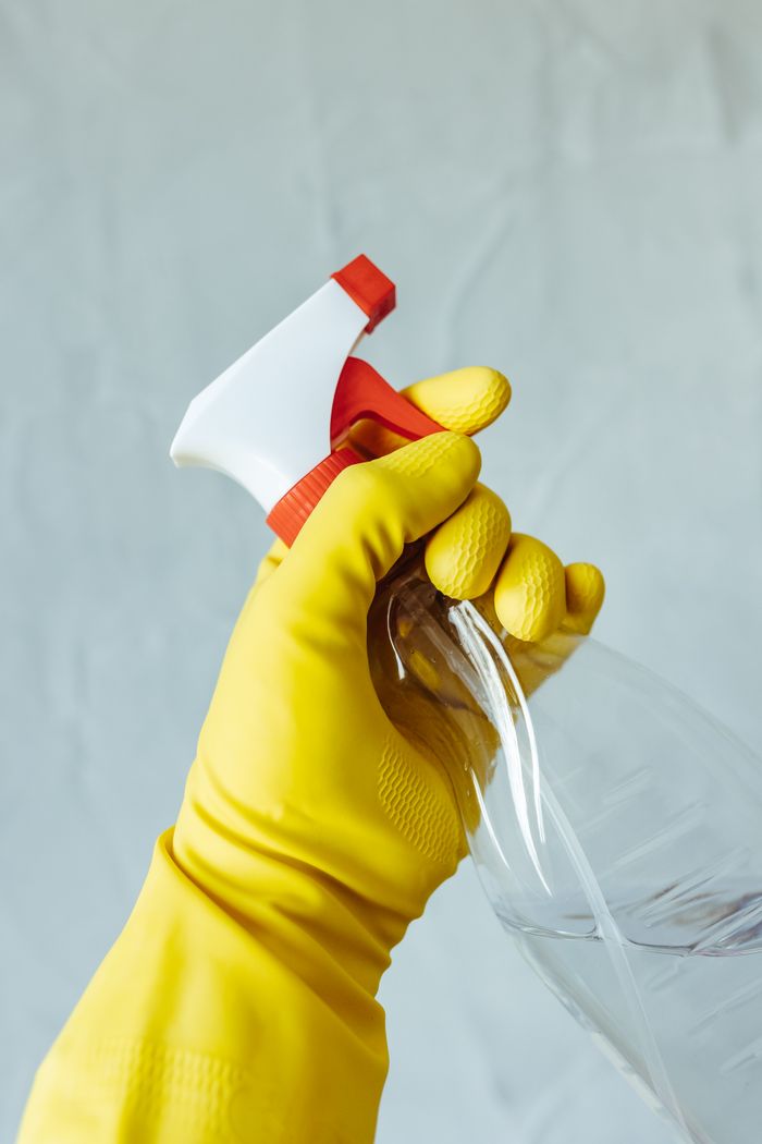 project-03-b A hand wearing a yellow rubber glove holds a red and white spray bottle against a neutral background.